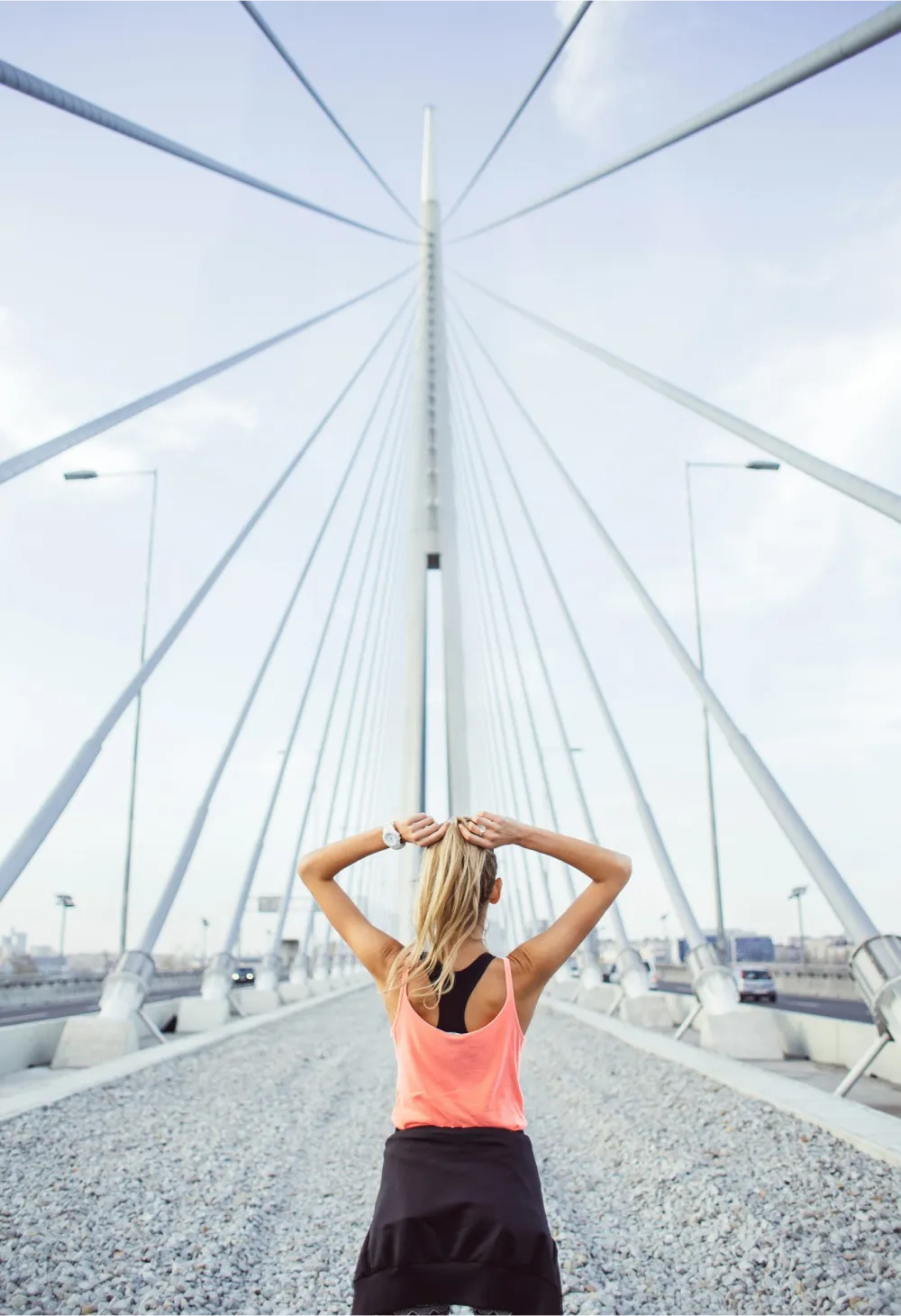 Photo of a woman wearing jogging athletic clothing. She's standing on the a bridge with her back facing the camera and her arms above her head.