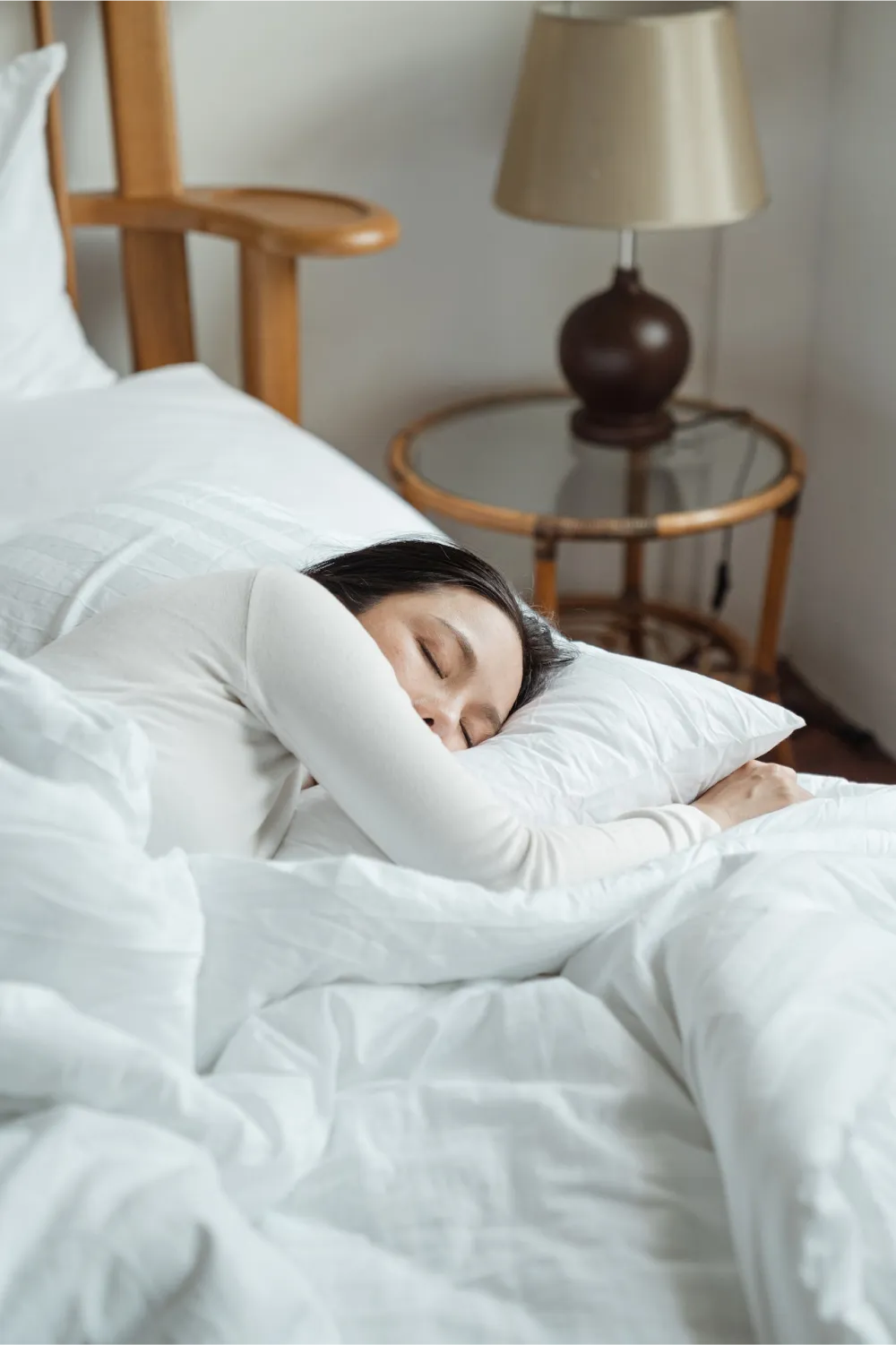 Photo of a woman sleeping peacefully in bed. The bed has white fluffy sheets with a night stand and a lamp in the top right corner of the image. 