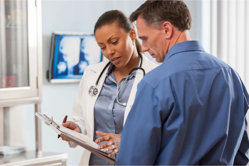 A doctor reading results to a patient off a clipboard. The doctor is is wearing a white lab coat and the patient is in a blue button down shirt.