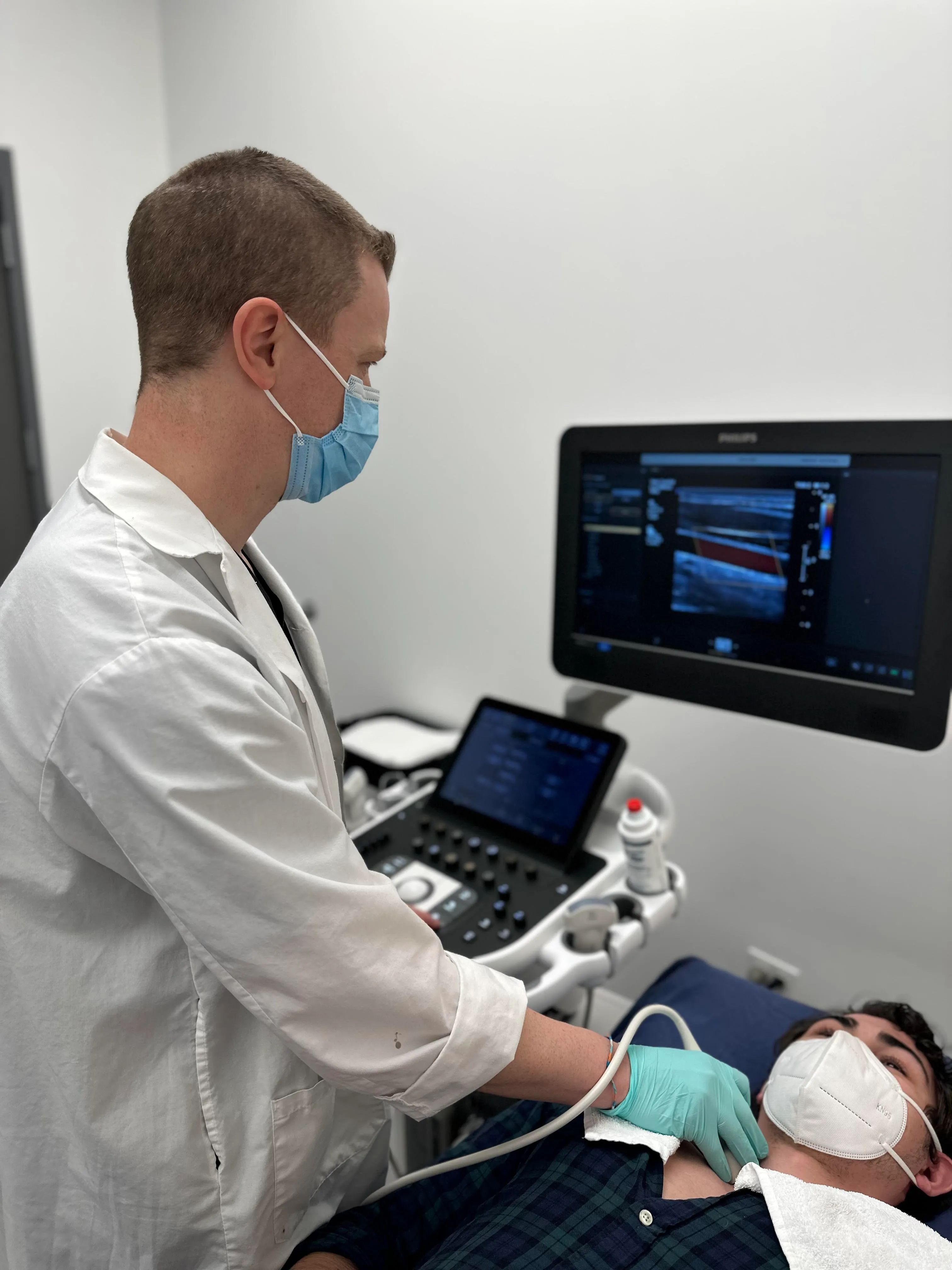 Technician performing an ultrasound treatment to a mans chest while he lays comfortably on an examination table. Both are wearing medical grade masks.